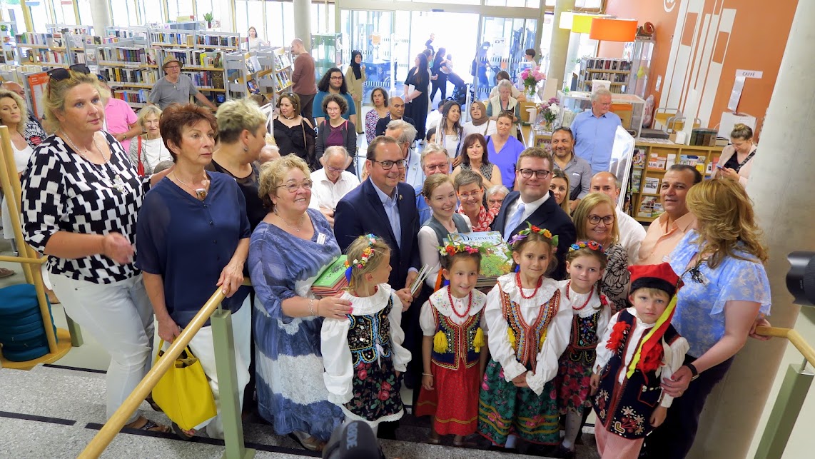 Tłum osób w różnym wieku zgromadzony w bibliotece. Uśmiechnięte dzieci w krakowskich strojach